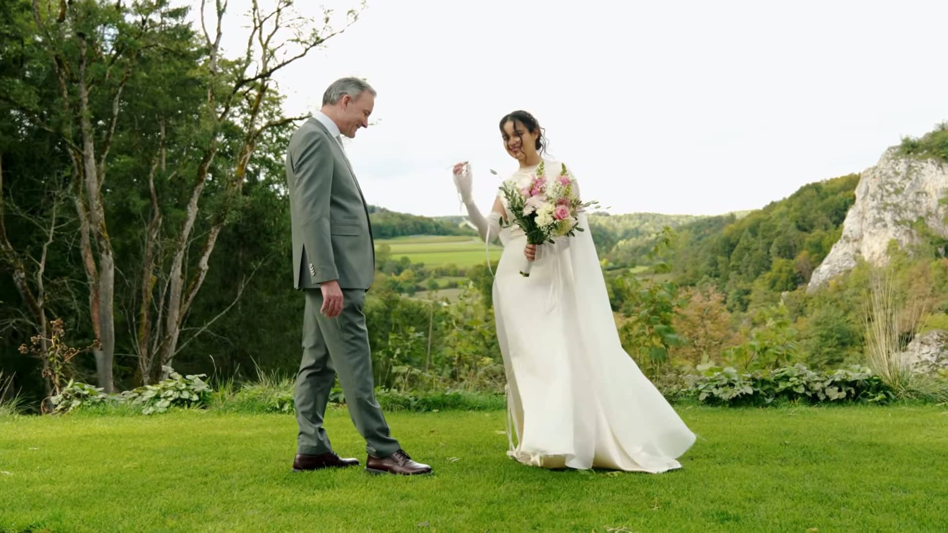 Ein Vater geht auf seine Tochter im Brautkleid zu, die auf einer Wiese über dem Tal auf ihn wartet.