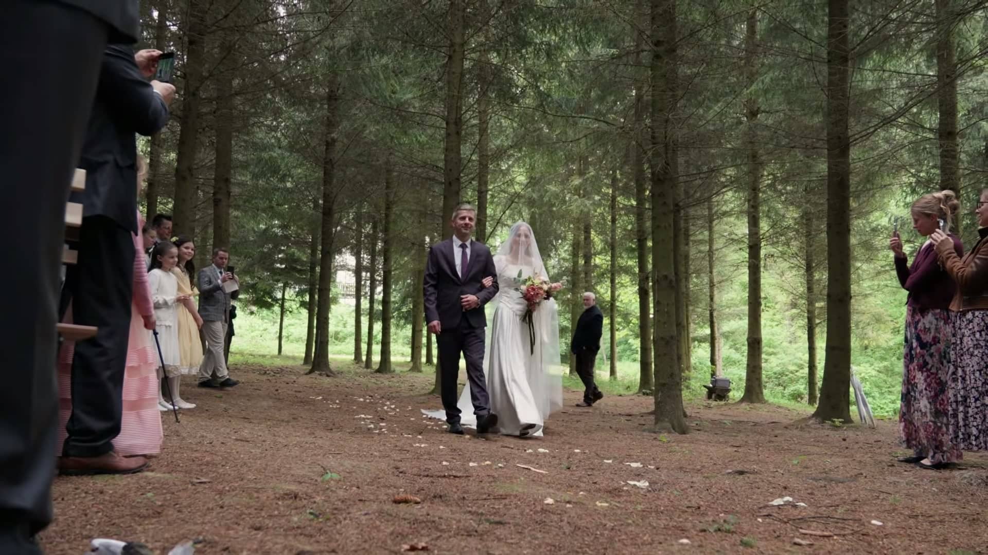 Der Vater führt die Braut zwischen hohen Fichten zur Trauung im Wald.